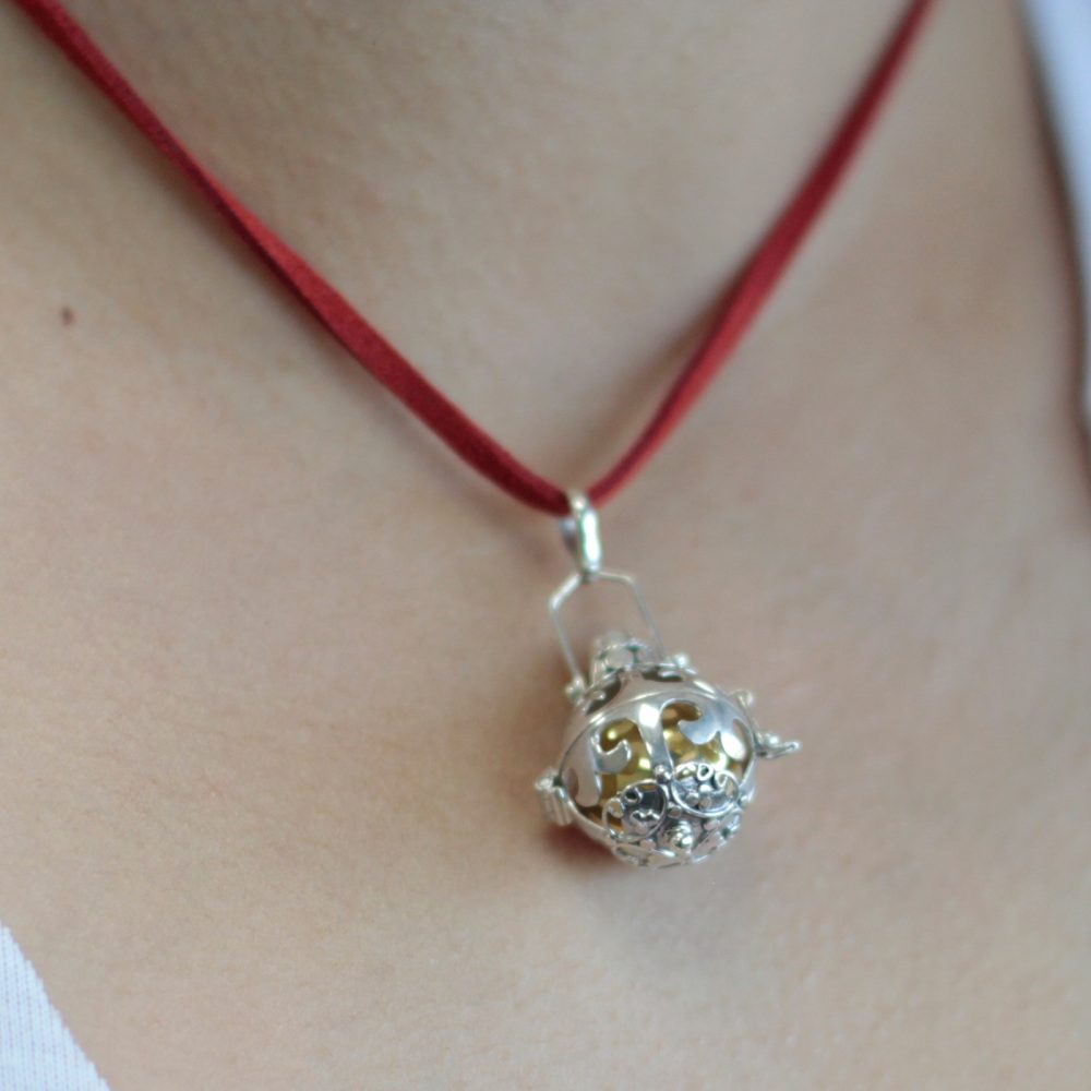 A silver filigree pendant hangs from a red cord around a person's neck. The pendant contains a golden ball inside its cage.