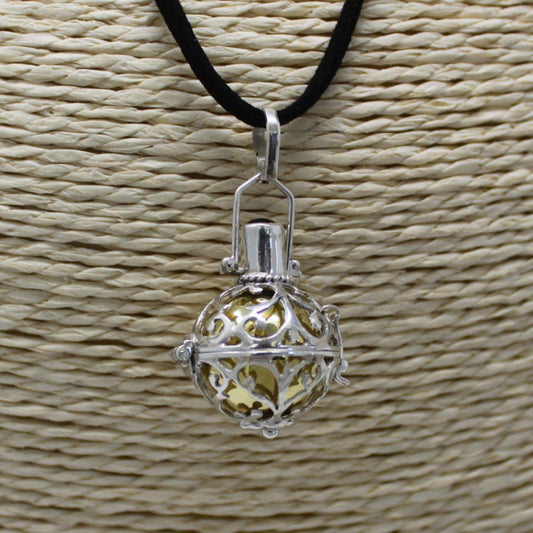 A silver, filigree pendant dangles from a black cord against a textured, neutral-colored background. The hollow pendant reveals a smooth, golden sphere inside.