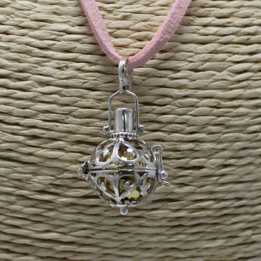 A silver pendant dangles from a pink cord against a woven mat. The pendant is spherical and ornately caged, revealing a gold interior.