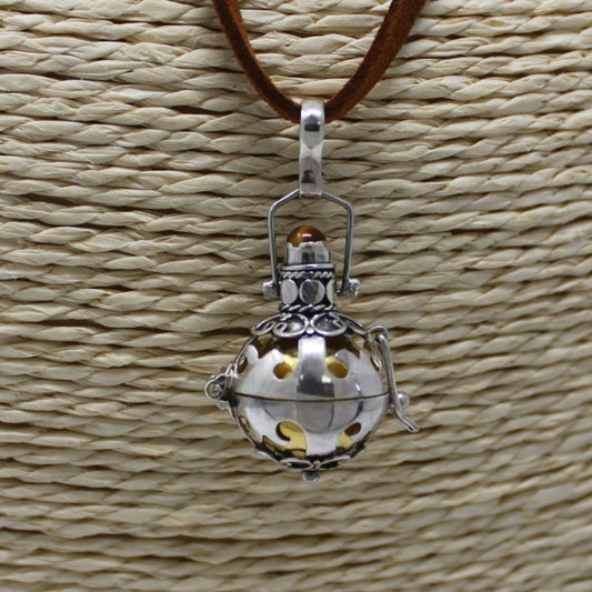 A silver pendant hangs from a brown cord against a textured beige background. The pendant is an ornate sphere with cut-out designs, revealing an inner golden hue.
