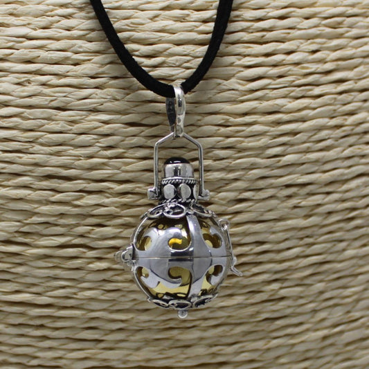 A silver, ornamental pendant dangles from a black cord. Its spherical, openwork design reveals a golden core, all displayed against a neutral woven background.