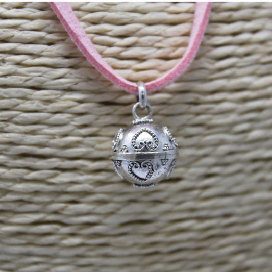 A silver, round pendant hangs from a pink, suede cord. The pendant is decorated with hearts, and it’s displayed against a textured, woven background.