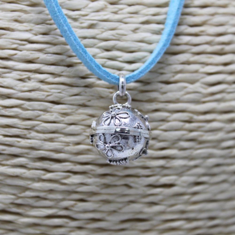 A silver pendant dangles from a blue suede cord. The pendant is a sphere decorated with flowers and scrollwork and is displayed against a background of tightly woven, tan material.