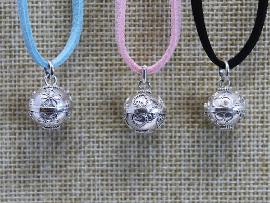 Three necklaces, each with a silver ball pendant, hang side-by-side. Each pendant features intricate floral or heart designs. The necklaces' cords are light blue, pink, and black, set against a woven background.