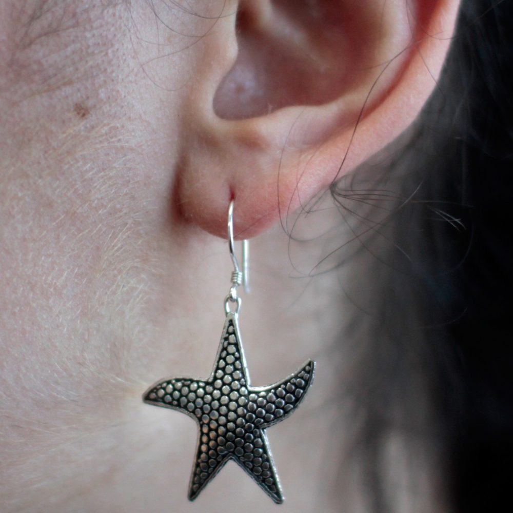 A starfish earring hangs from a pierced ear. The silver earring is textured with small bumps and is set against skin and dark hair.