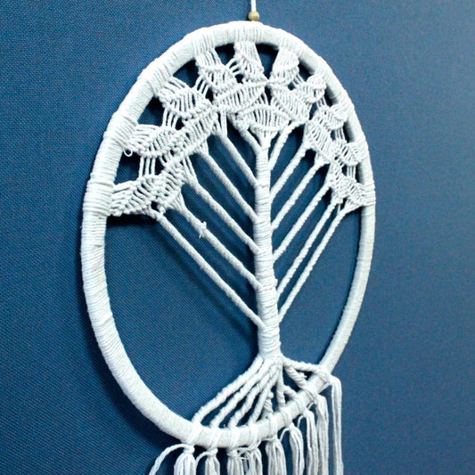 A white macrame tree hangs against a blue wall. The circular piece is made of knotted white cord and has a fringe at the bottom.