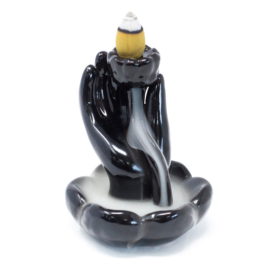 A black ceramic incense holder shaped like hands cradles a burning incense cone, with smoke cascading down into the petal-shaped base, on a white background.