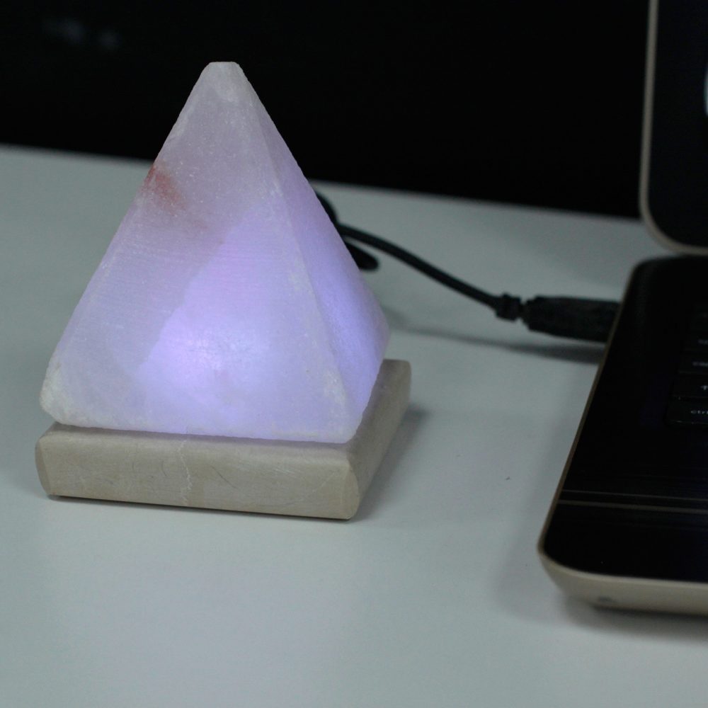 A glowing pyramid-shaped lamp emits a soft purple light on a desk, connected by a cable, next to an open laptop.
