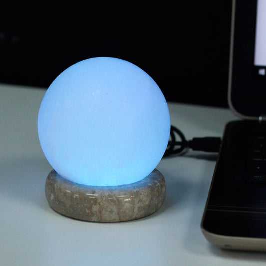 A glowing blue spherical lamp rests on a stone base, emitting light, connected to a laptop on a white table, surrounded by a dark background.