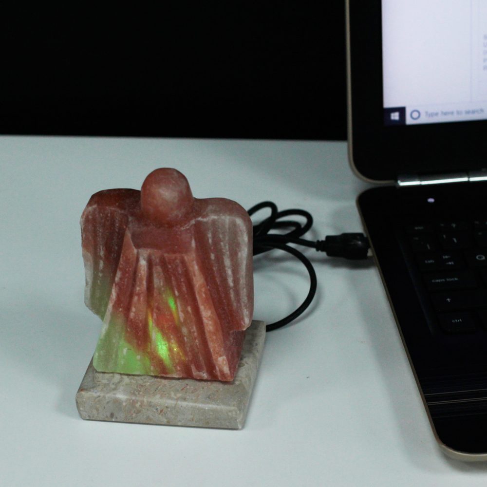 A red, abstract stone figurine with a glowing green light sits on a marble base, connected by a cable to an open laptop on a white surface.