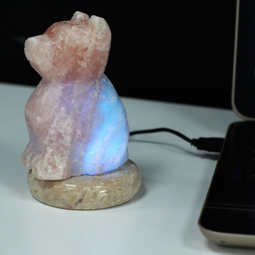 A salt lamp shaped like a bear emits a soft blue glow. It sits on a round base, connected via a USB cable, beside a laptop on a white surface.