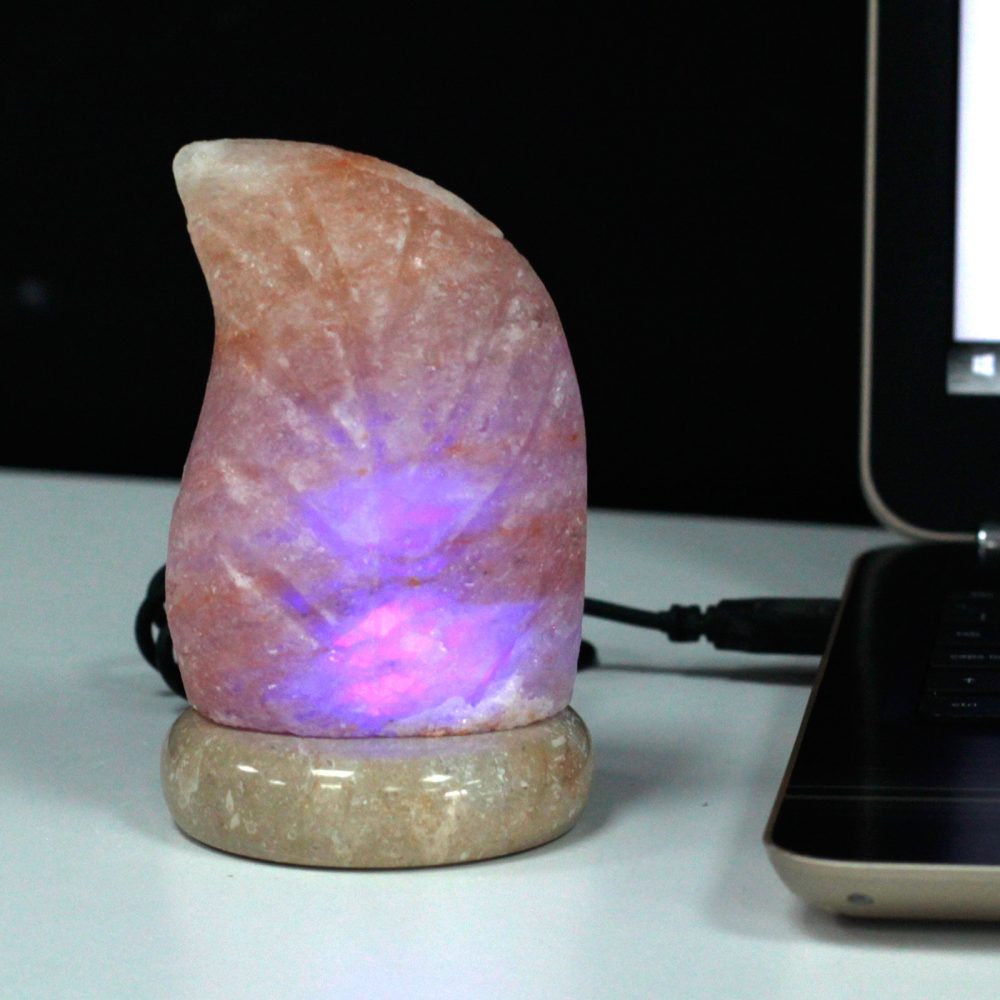 A pink salt lamp glows with purple light on a round base, placed on a white surface beside an open laptop.