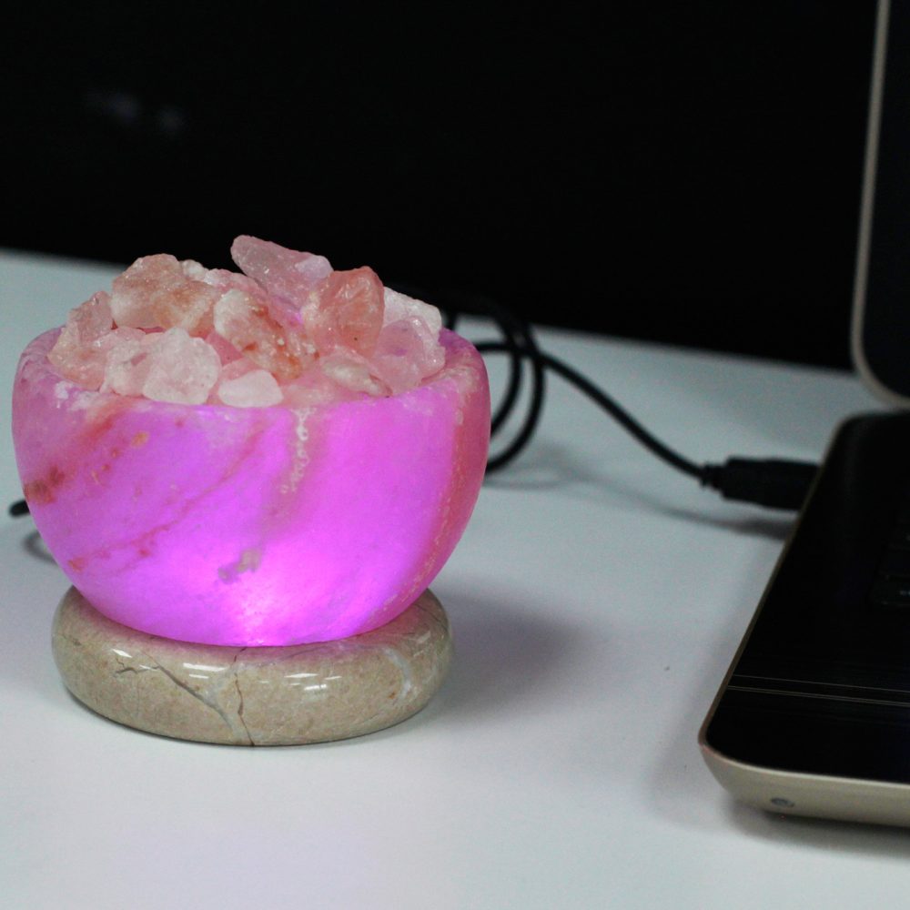 A pink, glowing salt lamp filled with pink crystals rests on a circular stone base, connected by a black cable. It sits on a white surface next to a partially visible laptop.