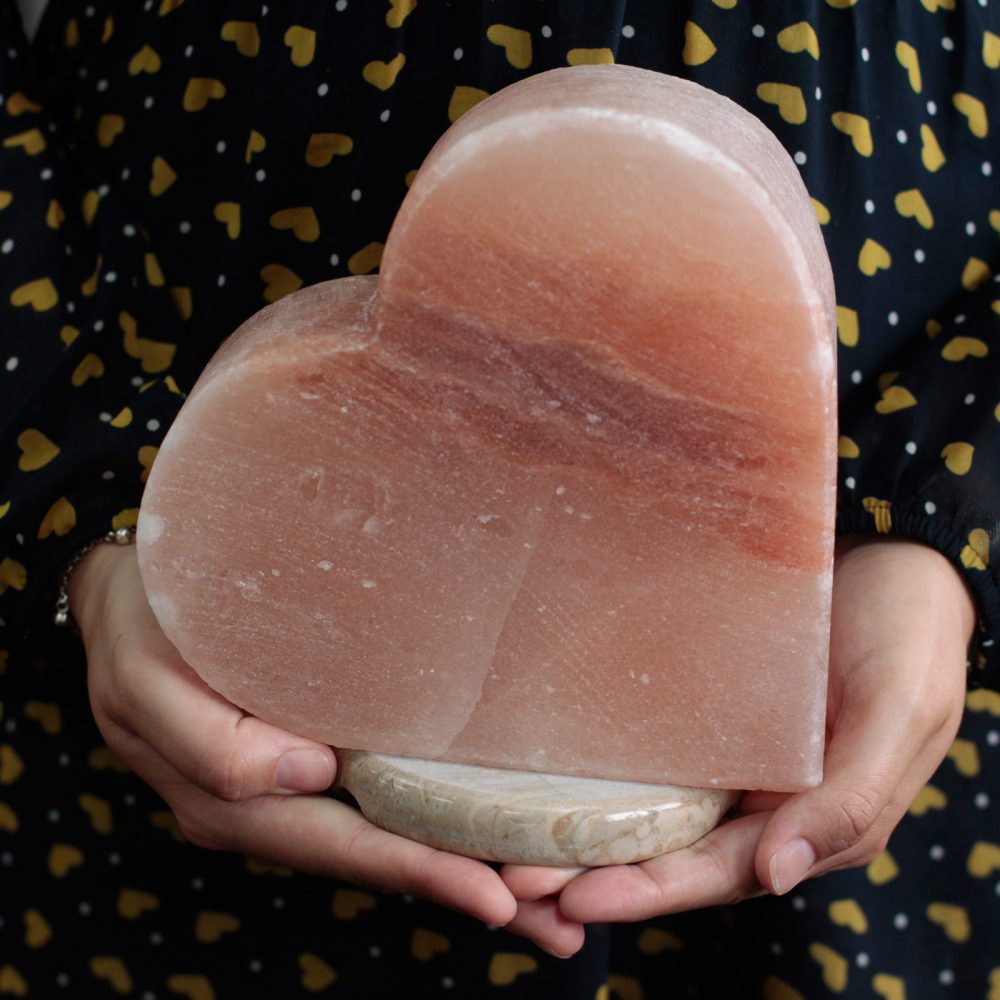 A person holds a large, heart-shaped, pink salt crystal with both hands. The individual wears a dark garment with yellow heart patterns.