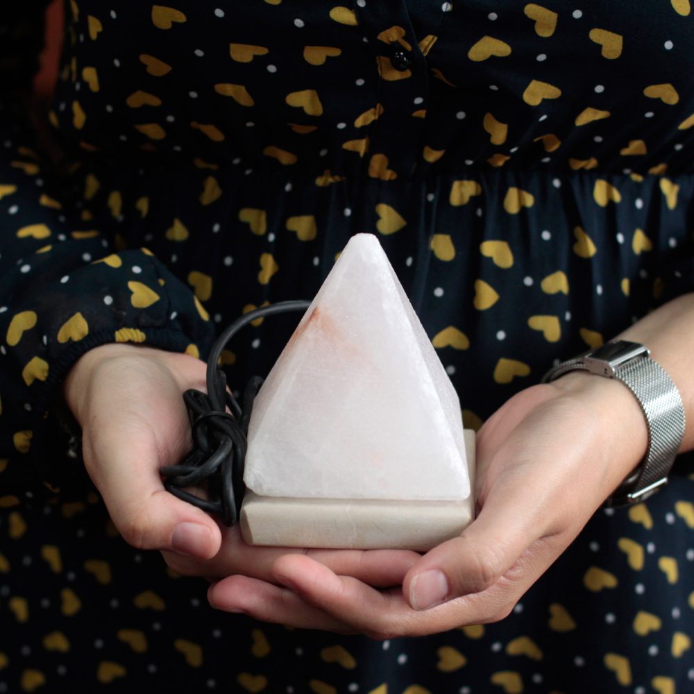 Hands hold a small, pyramid-shaped salt lamp with a black cord, set against a background featuring a dark dress with yellow heart patterns.