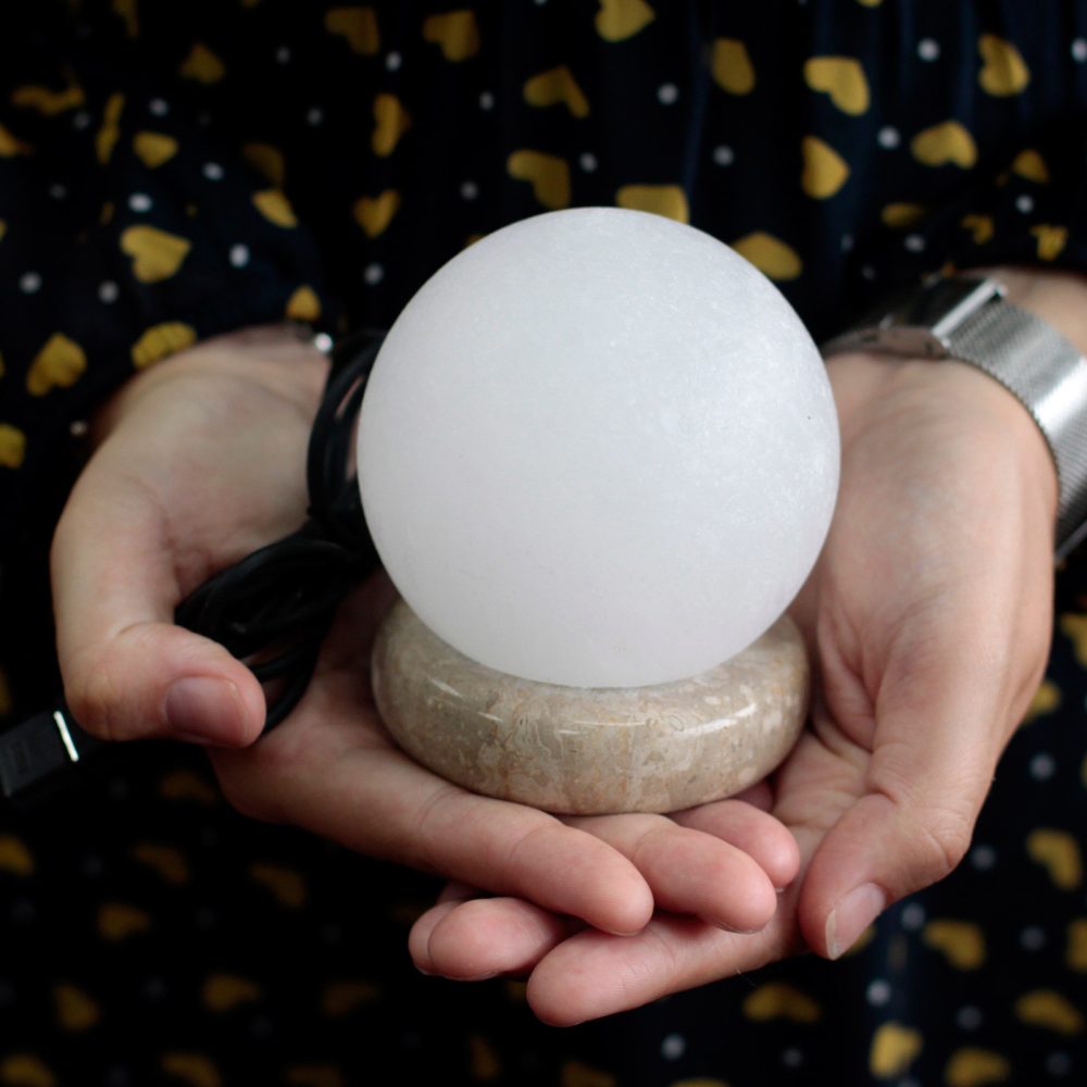 A white spherical lamp rests in hands, supported by a stone base, with a black cable extending from it. The person wears a polka-dotted garment and a silver wristwatch.