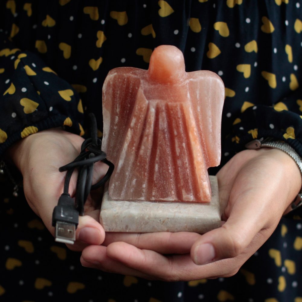 A person holds a carved pink salt lamp shaped like an angel with a USB cable, against a dark, heart-patterned background.