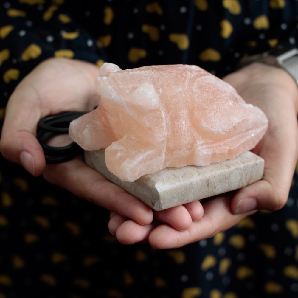 A pink, translucent salt crystal shaped like a pig rests on a small stone slab, held in two hands. Background shows a patterned fabric with yellow dots.