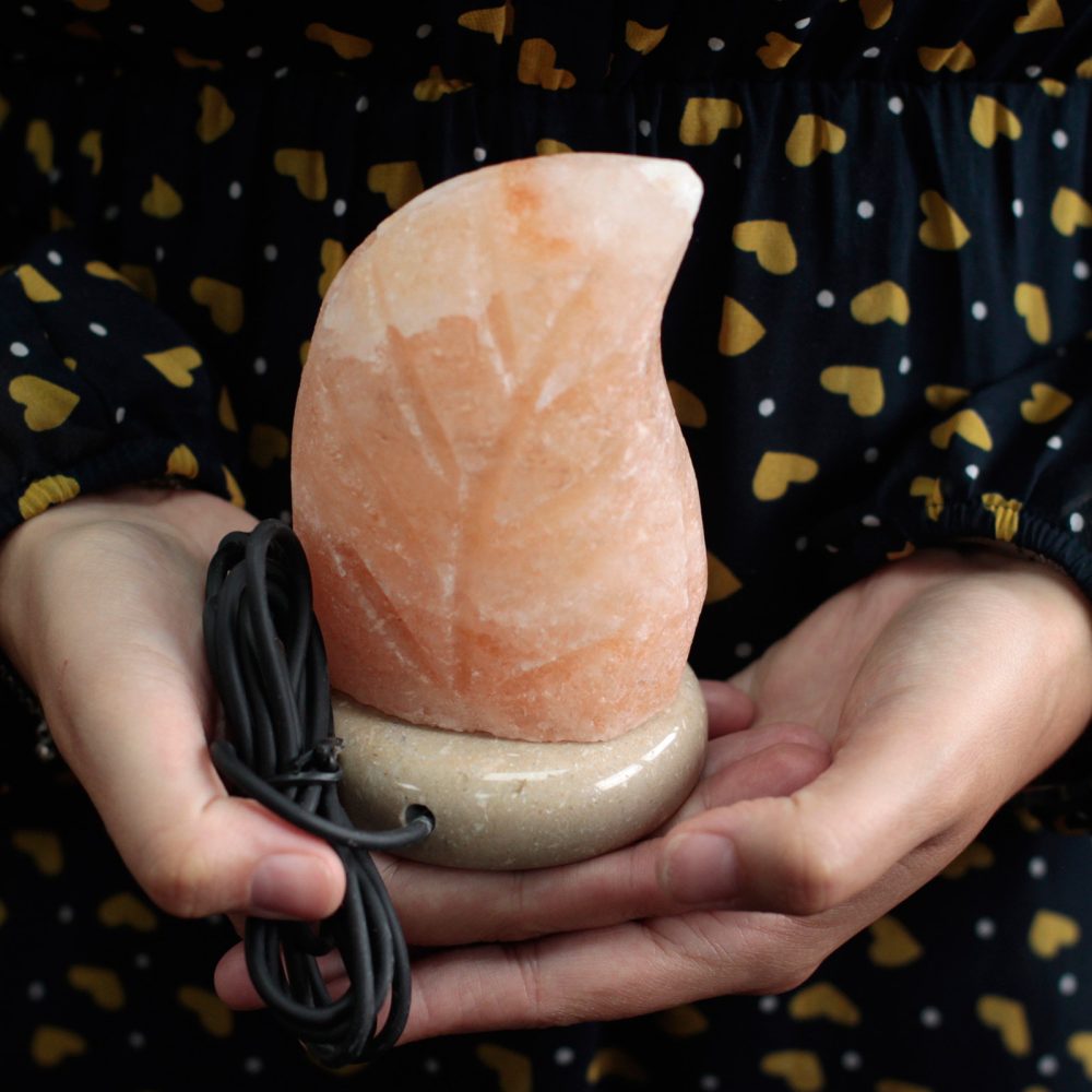A carved orange salt lamp rests in hands, with a black cord wrapped around it. The background is a dark fabric with yellow heart and white polka dot patterns.
