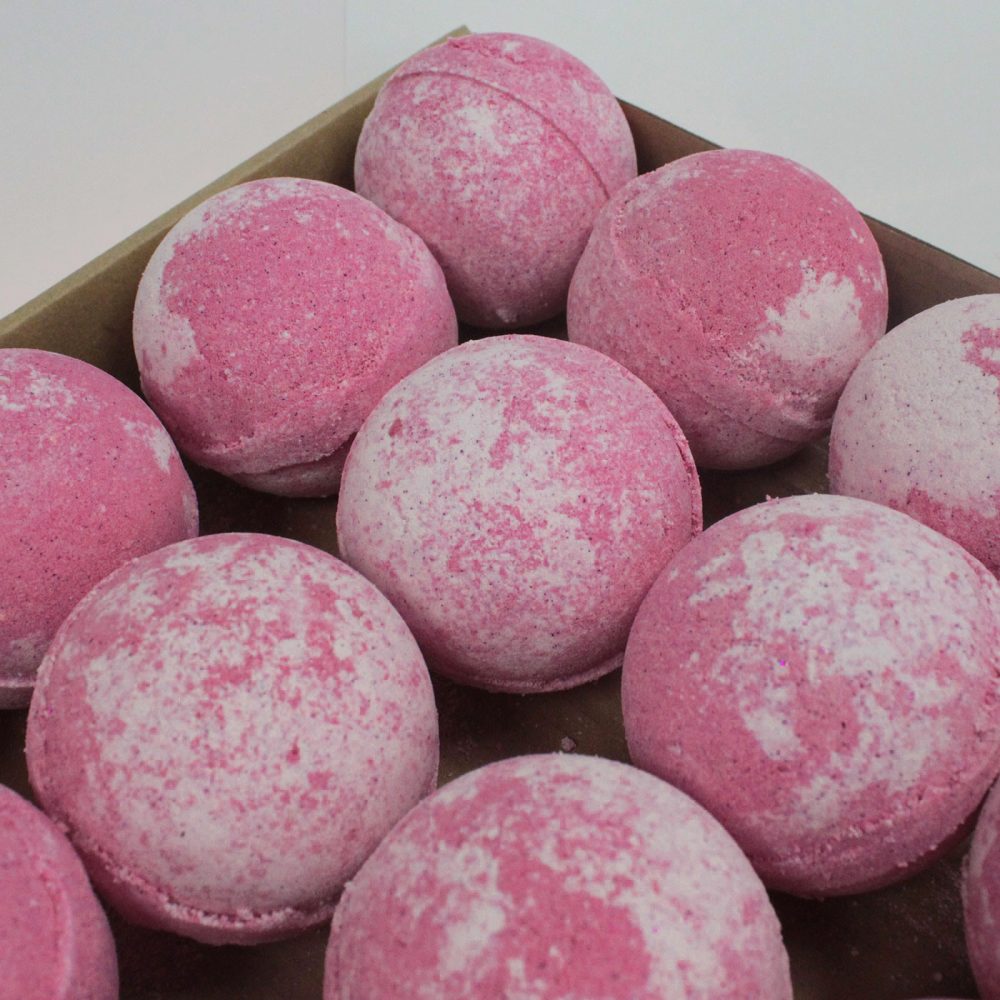 Pink and white bath bombs are arranged in a cardboard tray, each displaying a marbled pattern. The setting appears clean, highlighting the spherical shapes and vibrant colors.