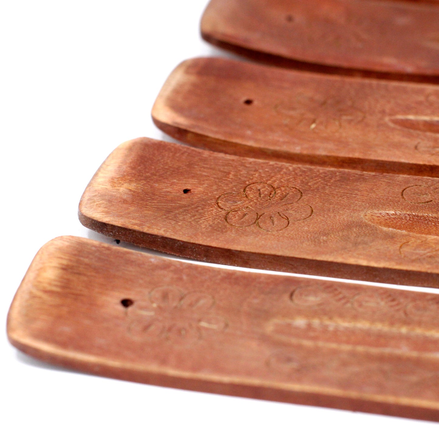 Wooden incense holders, engraved with floral patterns, are lined in a row. They have a smooth finish and small holes at one end, set against a plain white background.
