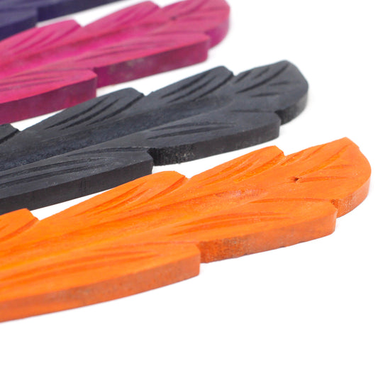 Colorful feather-shaped wooden pieces, in orange, black, and purple hues, lie flat in a row on a white surface, showcasing carved details and vibrant colors.
