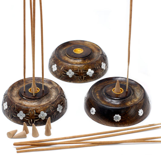 Three wooden incense holders feature floral decorations, holding incense sticks and cones. The setup includes additional incense sticks and cones on a white surface, suggesting a peaceful, aromatic setting.