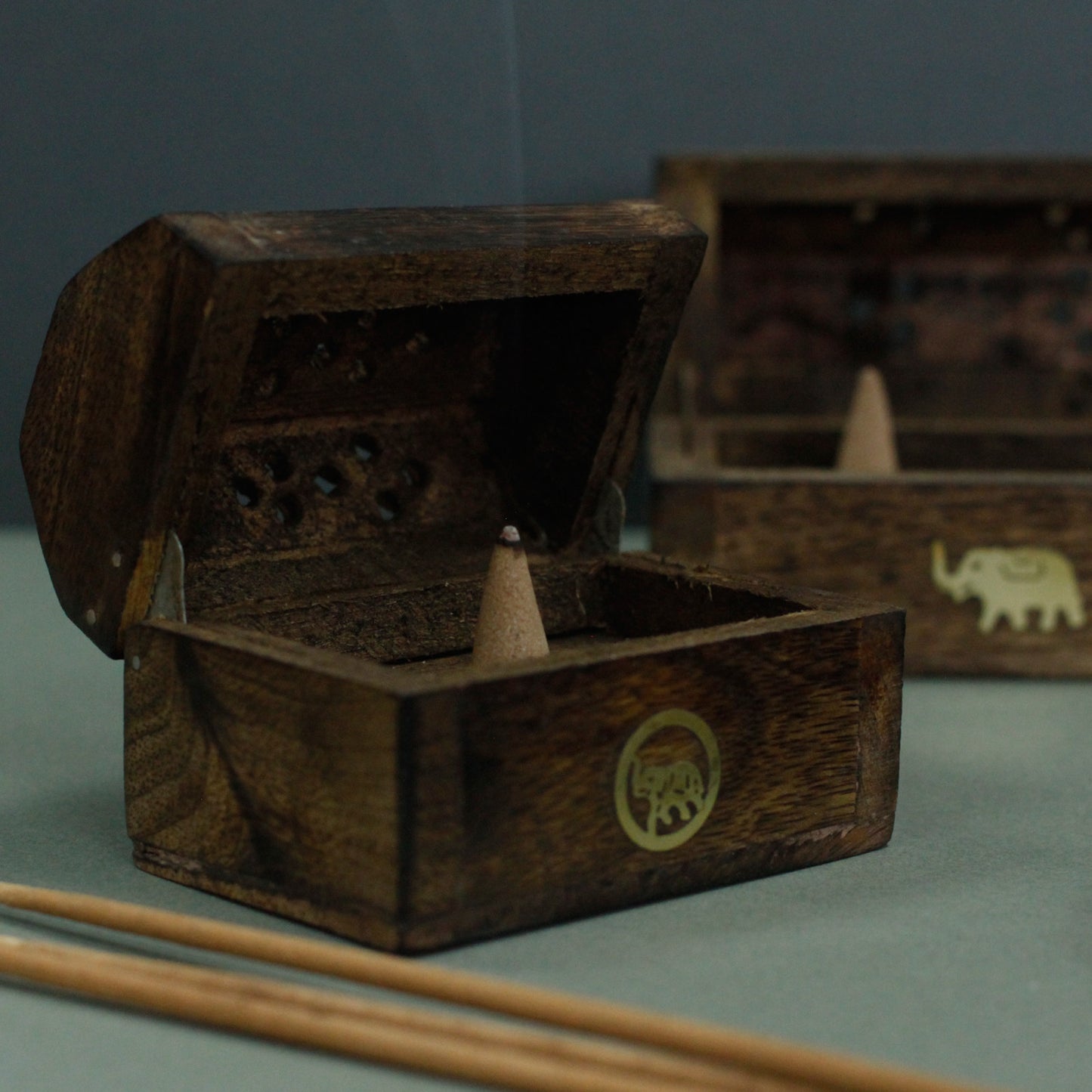 A wooden incense box with a carved elephant design releases smoke from a burning incense cone inside. Additional boxes are visible in the background on a dark surface.