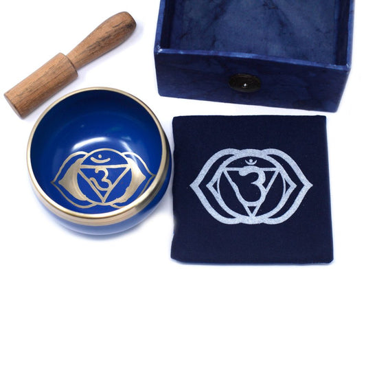 A blue singing bowl adorned with a golden chakra symbol lies alongside a wooden striker, a matching cloth with the same symbol, and an empty blue square box.