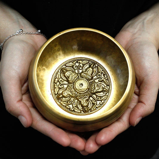 A brass bowl with intricate designs inside is cradled by two hands. The background is dark, highlighting the detailed patterns and smooth, polished surface of the bowl.