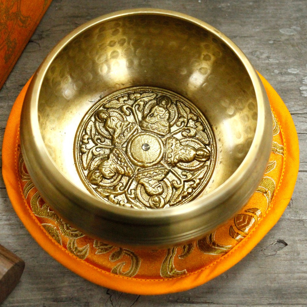 A golden singing bowl rests on an ornate orange cushion. The intricate design inside features symmetrical patterns. The setting is a wooden surface, giving a rustic ambiance.