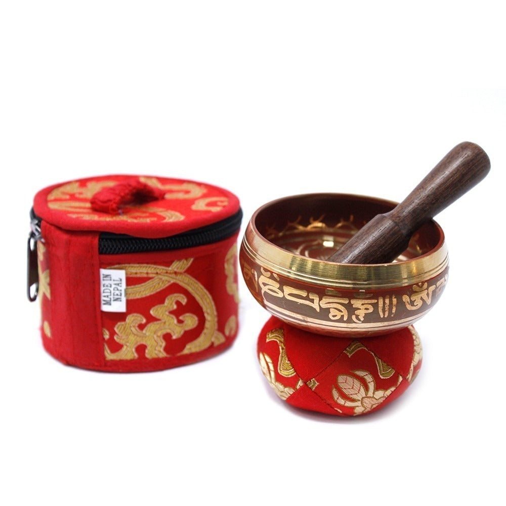 A Tibetan singing bowl with a wooden striker rests on a decorative red cushion. Nearby, a matching red case labeled "Made in Nepal" features gold patterns, enhancing the traditional setup.