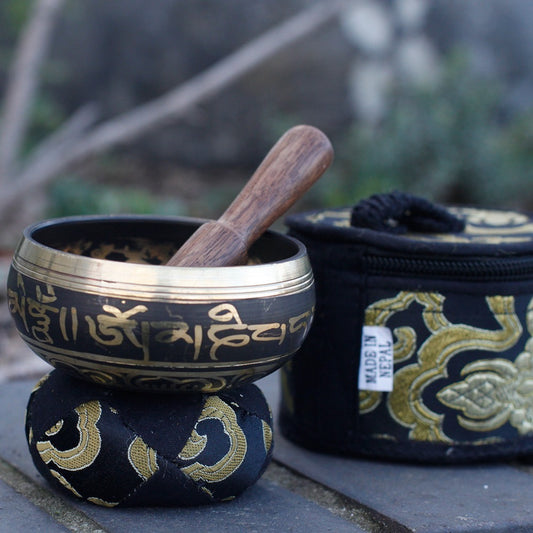 A singing bowl rests on an embroidered cushion, with a wooden mallet inside. Beside it, a matching pouch labeled "MADE IN NEPAL." The background features blurred greenery.