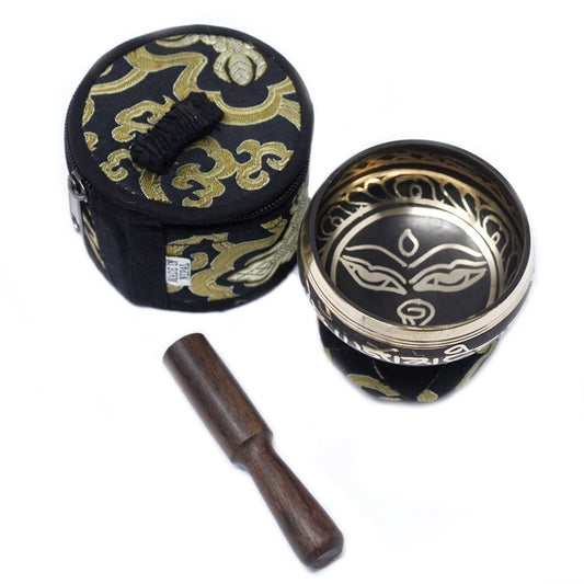 A singing bowl with intricate designs rests beside an ornate fabric case and a wooden striker, set against a plain background. The bowl features detailed patterns and script inside.