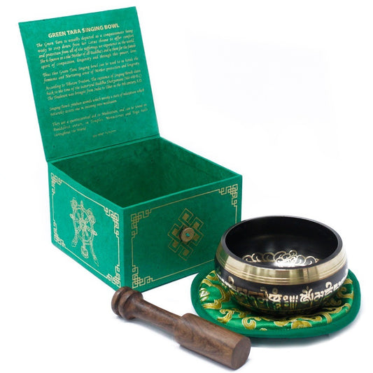 A carved singing bowl rests on a decorative cushion beside a wooden mallet. Nearby, a green box lid displays text: "GREEN TARA SINGING BOWL," detailing its symbolism and uses in meditation.