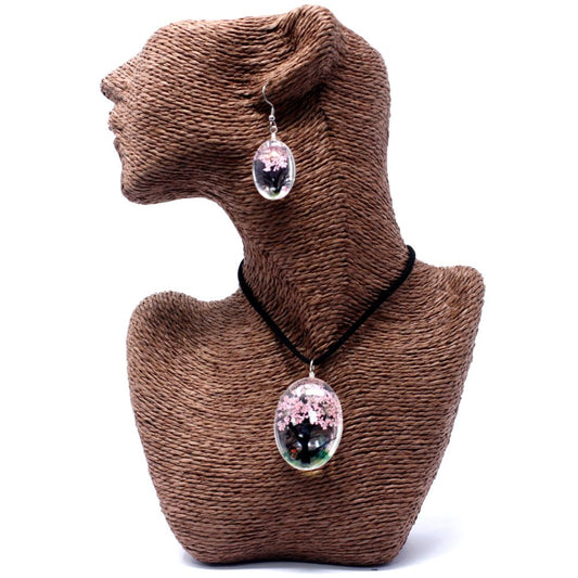 A brown textured mannequin displays a matching necklace and earring set, each containing pink blossom inclusions within a clear, oval resin pendant, against a stark white background.