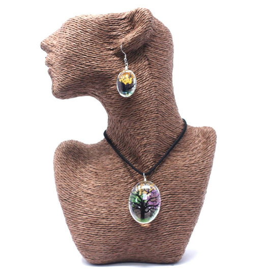 A tan textured mannequin displays an oval pendant with a colorful tree design on a black cord, and a matching earring, against a white background.
