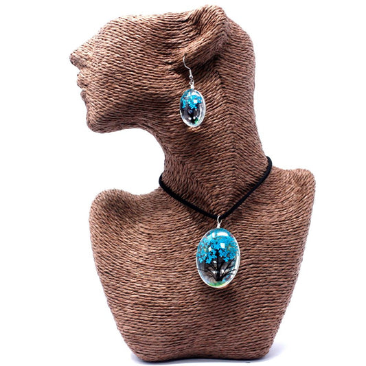 A brown mannequin displays jewelry. A necklace with a blue tree pendant sits on its chest, and a matching earring is on its ear. They are on a white surface.