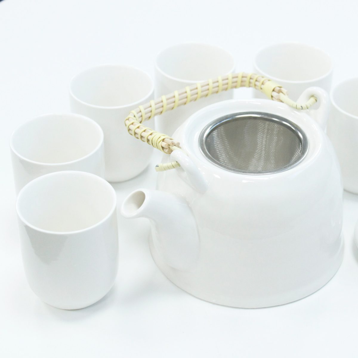 A white teapot with a woven handle is surrounded by six matching cups on a plain white surface, creating a minimalist and serene setting.