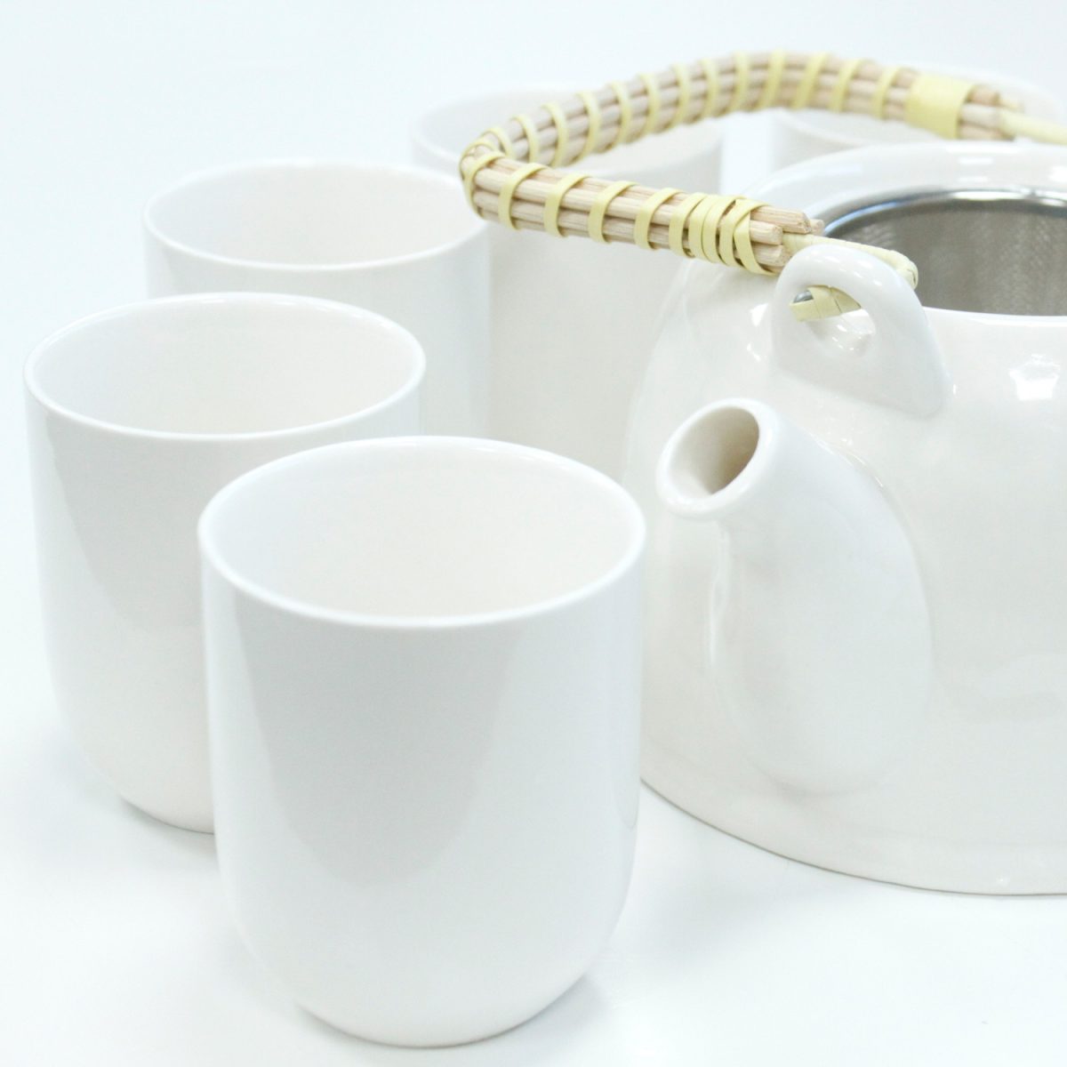 White ceramic teapot with a woven handle stands beside four matching cups on a smooth, white surface.
