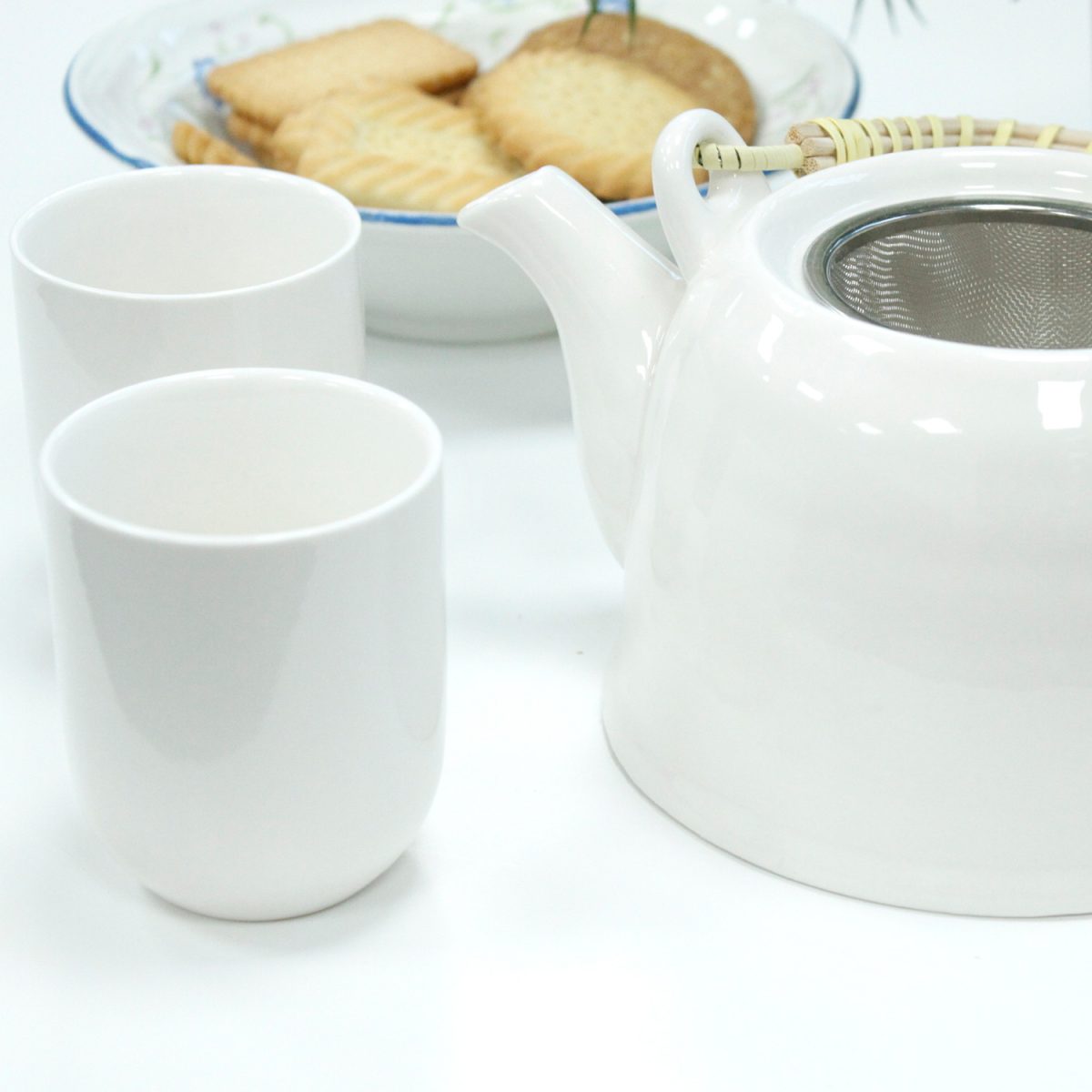 White teapot with bamboo handle sits beside two white cups, while a plate of cookies is slightly blurred in the background on a white surface.