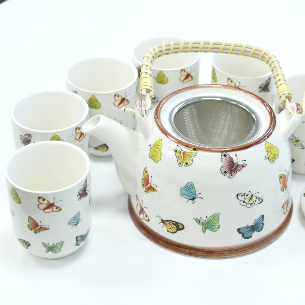 A ceramic teapot with a wicker handle, featuring colorful butterfly designs, sits surrounded by six matching cups on a white surface.