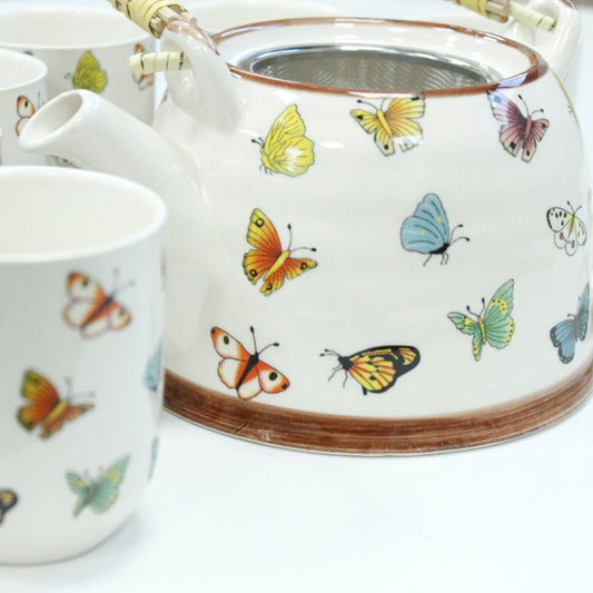A ceramic teapot and cups adorned with vibrant butterfly illustrations are set on a white surface. The teapot features a mesh filter and woven handle accents.