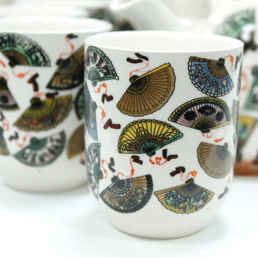 Ceramic mugs feature intricate fan designs in various colors, including green, blue, and yellow, against a white background. The mugs are placed on a light surface, creating a vibrant display.