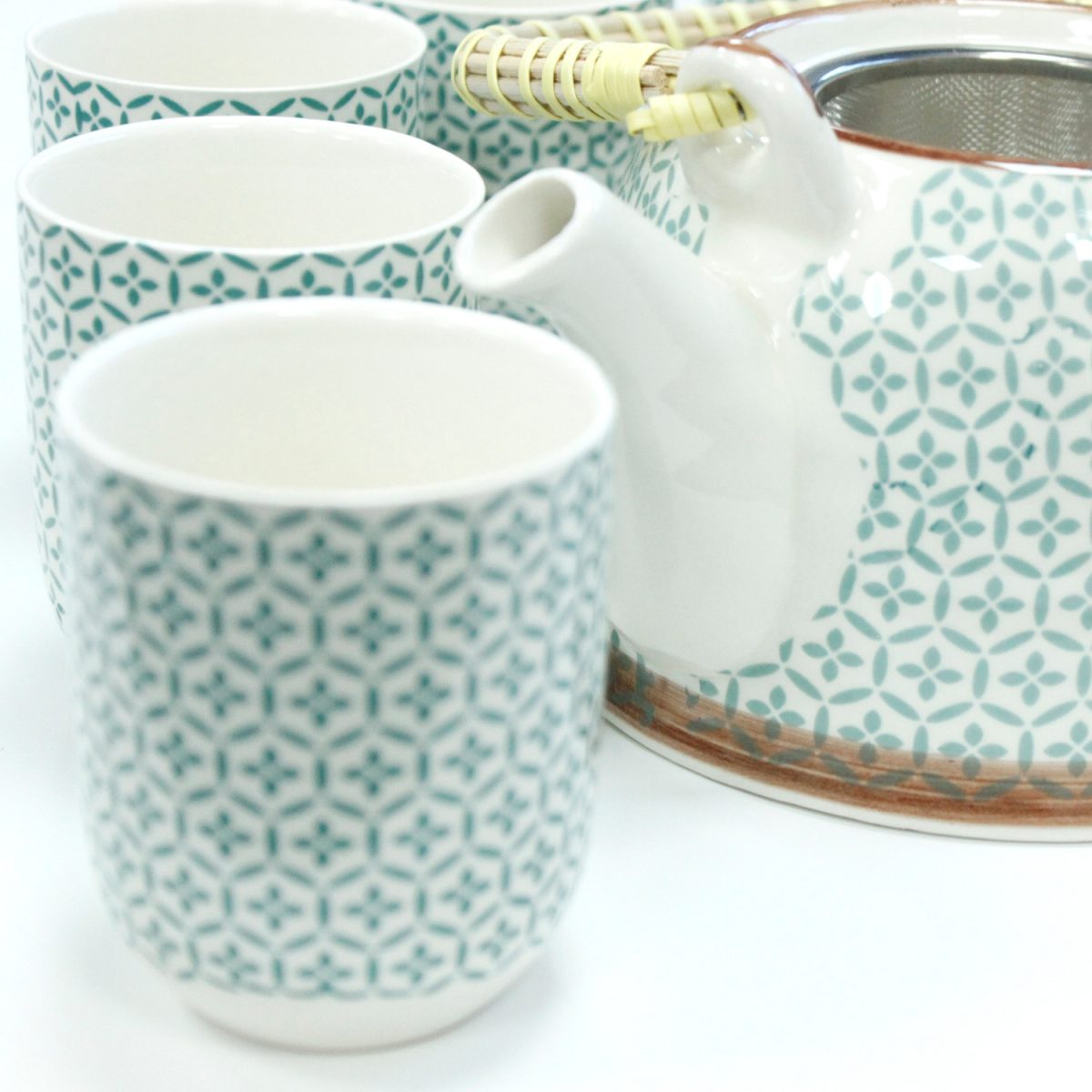 Teapot with a wicker handle and three cups, all decorated with a teal geometric pattern, sit on a white surface.