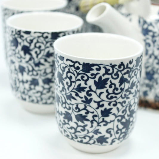 Ceramic cups feature intricate blue floral patterns on a white background. They are positioned closely together, accompanied by a partially visible teapot in a similar style, set against a plain, light backdrop.