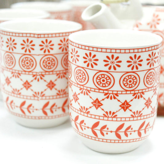 Mugs feature red geometric and floral patterns, including circles, stars, and leaves, on a white ceramic surface. Other similar mugs and objects surround them.