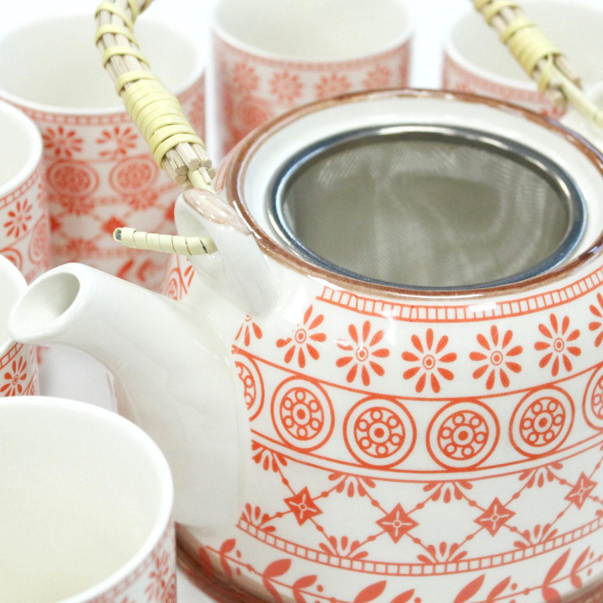 Ceramic teapot with orange floral and geometric patterns, featuring a bamboo handle and surrounded by matching cups, set in a bright, minimalist environment.
