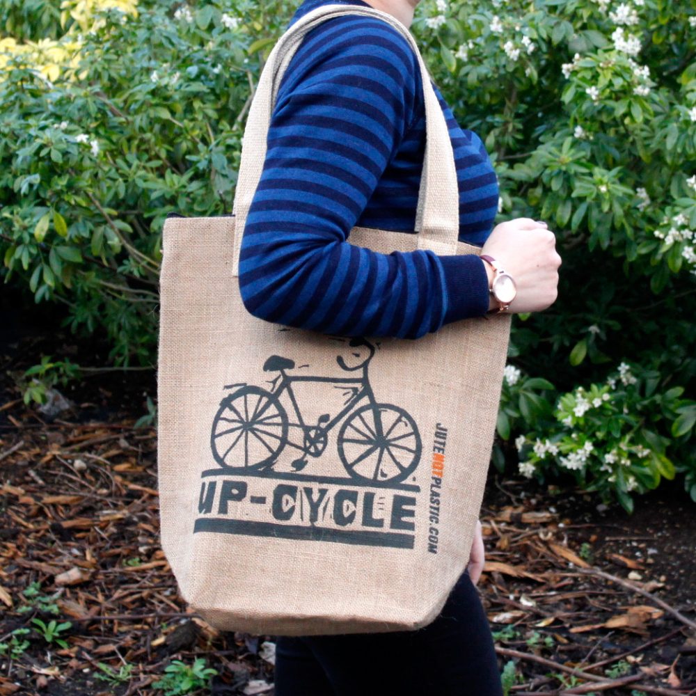 A person holds a tan tote bag that displays a black bicycle graphic and the words "UP-CYCLE JUTENOTPLASTIC.COM" while standing outside amongst foliage.