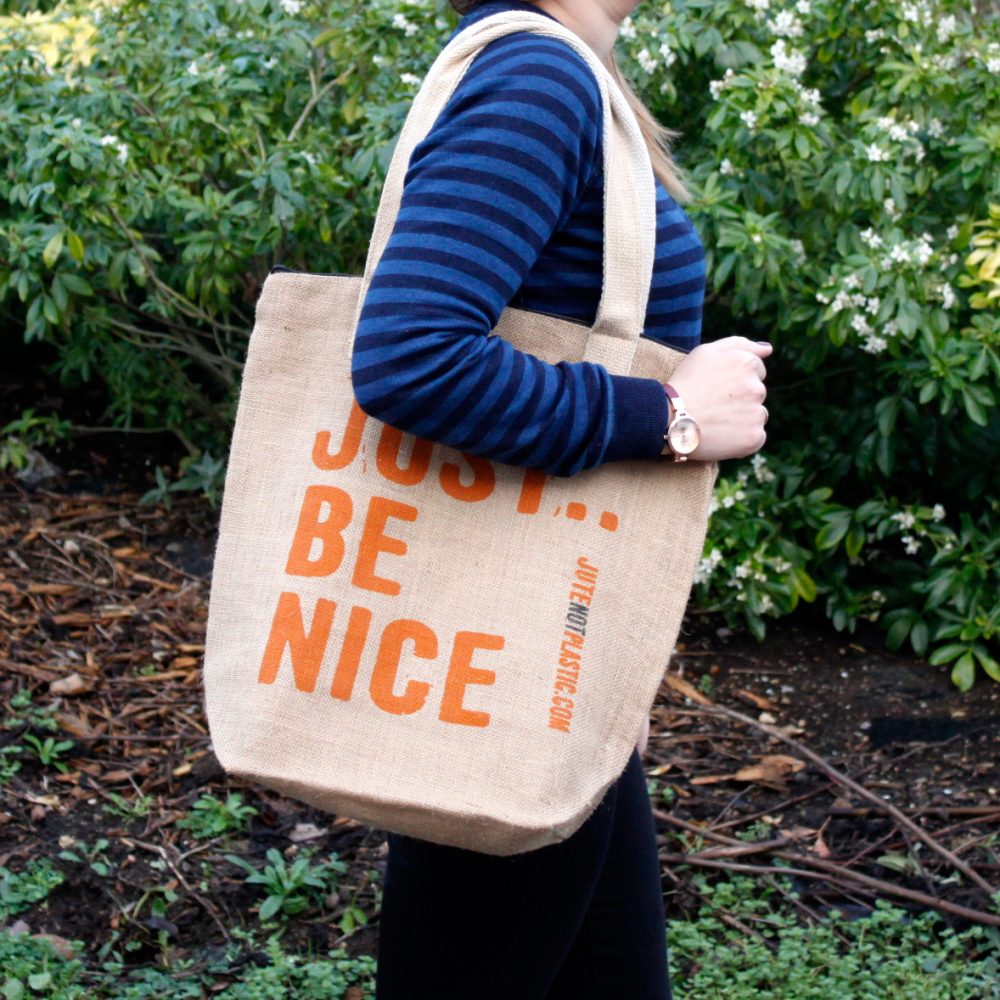 A tan tote bag reading "JUST BE NICE JUTENOTPLASTIC.COM" hangs on a woman's shoulder, with greenery behind her.